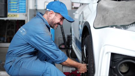 Photo for Automotive mechanic man in uniform with screwdriver repairing car wheel and checking tire at vehicle repair shop. Car service, repair, maintenance and man mechanic concept - Royalty Free Image