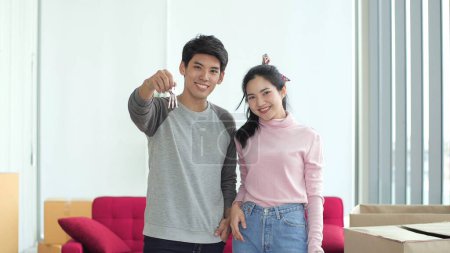 Téléchargez les photos : Portrait de jeune couple asiatique debout et tenant la clé de la maison pour la nouvelle maison et regardant vers la caméra. Ils ont acheté une maison ou un appartement neuf - en image libre de droit