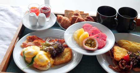 Photo for Breakfast on bed served with cup of coffee, fresh fruits and eggs benedict, croissant on wooden tray. Room Service in Hotel - Royalty Free Image