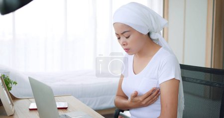 Foto de Mujer asiática con cáncer y pañuelo en la cabeza tiene dolor en el pecho mientras trabaja con la computadora portátil en casa. Problemas respiratorios. Sufrimiento de dolor en el pecho - Imagen libre de derechos