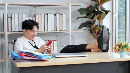 Foto de Joven asiático hombre poniendo los pies en la mesa en la oficina, mientras que el uso de teléfono inteligente disfrutar de los mensajes de chat en las redes sociales, ver contenido de Internet tomar un descanso del trabajo - Imagen libre de derechos