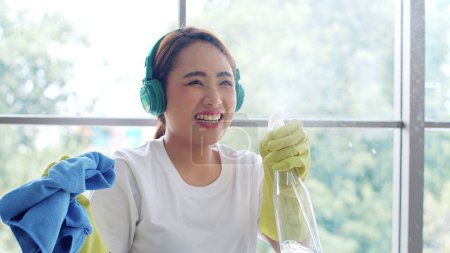 Foto de Feliz joven asiática mujer con auriculares y guantes de goma sosteniendo spray de limpieza y trapo mientras limpia ventanas en casa. Trabajador doméstico y concepto de limpieza - Imagen libre de derechos