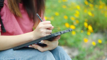 Téléchargez les photos : Fermez les mains de l'adolescente étudiante écrivant et utilisant un stylo avec une tablette numérique à l'extérieur. Dessinez ou connectez-vous sur l'écran de tablette numérique vierge. Concept technologique - en image libre de droit