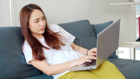 Foto de Mujer asiática joven usando el ordenador portátil mientras está acostado en el sofá en la sala de estar. Trabajando desde casa durante. Mujer acostada en el sofá escribiendo en el ordenador portátil - Imagen libre de derechos
