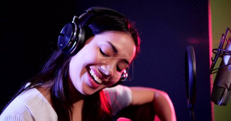 Téléchargez les photos : Heureuse jeune femme asiatique portant un casque jouant de la guitare enregistrement acoustique en studio. Femme exécutant guitare acoustique solo intro - en image libre de droit