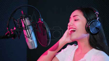 Foto de Joven mujer asiática artista cantando canción en estudio de música. Mujer cantante con auriculares usando micrófono cantando una canción en estudio de neón de grabación - Imagen libre de derechos