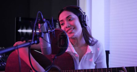 Photo for Young asian woman performing guitar acoustic solo intro in recording songs in studio.  woman artist wearing headphones playing guitar acoustic recording in neon studio - Royalty Free Image