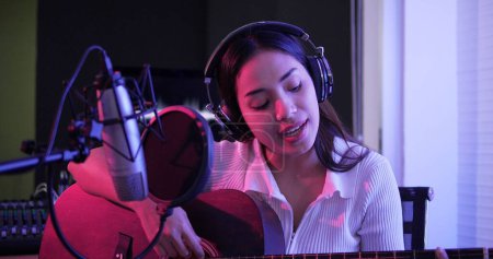 Foto de Mujer asiática joven tocando guitarra acústica solo intro en la grabación de canciones en el estudio. mujer artista con auriculares tocando la guitarra grabación acústica en estudio de neón - Imagen libre de derechos