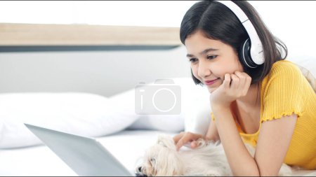Téléchargez les photos : Heureuse fille asiatique dans les écouteurs sans fil modernes couché et se détendre sur le lit avec chien tout en blessant film par ordinateur portable le week-end à la maison. Jeune femme dans les écouteurs - en image libre de droit