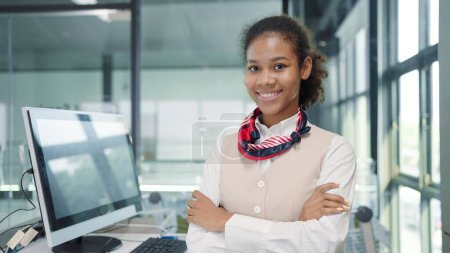 Téléchargez les photos : Gros plan visage souriant de jeunes femmes africaines américaines hôtesse de l'air sur le bureau au sol un service debout avec les bras croisés regardant la caméra - en image libre de droit