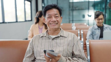 Téléchargez les photos : Gros plan Visage souriant d'un homme asiatique âgé assis à la porte de l'aérogare de l'aéroport, tenant un smartphone et un billet d'embarquement tout en attendant un vol d'avion regardant une caméra. Concept de voyage - en image libre de droit