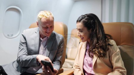Foto de Dos personas de negocios pasajeros en el asiento en el avión mirando juntos el teléfono inteligente, comprobando el correo electrónico o plan de viaje de negocios. Disfrutando de un largo vuelo de negocios. Viaje de negocios, concepto de tecnología - Imagen libre de derechos