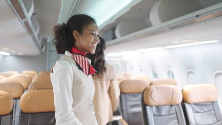 Foto de Atractiva azafata afroamericana en uniforme de pie y sonrisa en la clase económica del avión. Mujer tripulación de cabina saludando a los pasajeros en el servicio de avión. Concepto de vías aéreas - Imagen libre de derechos