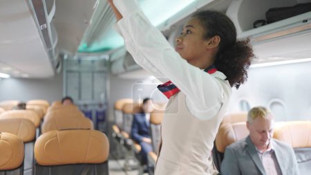 Photo for African american women flight attendant in uniform closing overhead luggage bin in airplane. Checking overhead luggage bin before airplane take off - Royalty Free Image