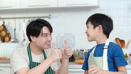 Téléchargez les photos : Heureux fier jeune père asiatique montrant pouce jusqu'à petit fils. joyeux asiatique père donnant pouces jusqu'à petit fils dans cuisine heureux asiatique famille concept - en image libre de droit