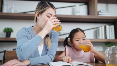 Foto de Alegre madre asiática e hija pequeña disfrutan bebiendo jugo de naranja juntos en la sala de estar en casa. Concepto de relación madre-hijo - Imagen libre de derechos
