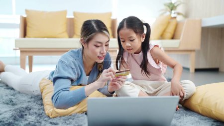 Photo for Happy asian mother and little daughter holding credit card for making online shopping on laptop while lying on floor in living room enjoying leisure together. Online shopping concept - Royalty Free Image