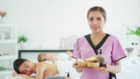 Foto de Mujer asiática masajista profesional en bandeja de sujeción uniforme con suministros de spa sonriendo a la cámara mientras está de pie en el spa y sala de masajes. Bienestar, salud y masaje - Imagen libre de derechos