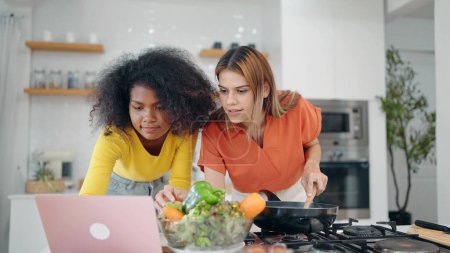 Foto de Dos amigas jóvenes usando labtop mientras preparan comida juntas en la cocina de su casa. Mujer lesbiana pareja aprendiendo a cocinar de video clip en línea en labtop. Vida LGBT, Pareja lesbiana - Imagen libre de derechos