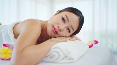 Foto de De cerca la cara de la hermosa joven asiática en toalla tumbada en la cama de spa mirando a la cámara mientras se relaja en vacaciones en el salón de spa. Tratamiento de spa, concepto de cuidado corporal - Imagen libre de derechos