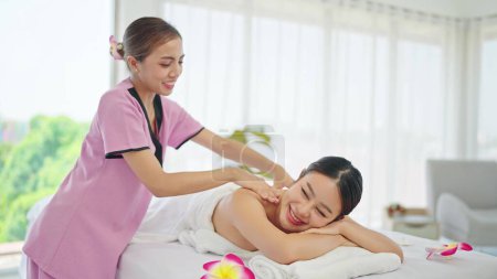 Téléchargez les photos : Femme asiatique allongée sur un lit de massage tout en relaxant massage du dos par une thérapeute au salon de soins de spa de beauté. Masseuse asiatique femme parler avec femme client tout en massant retour - en image libre de droit