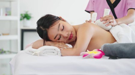 Téléchargez les photos : Belle femme en serviette couchée sur un lit de spa se faire masser avec des herbes boule chaude par masseur thérapeute au salon de spa. Thérapie, Massage thaïlandais, Soins du corps concept - en image libre de droit
