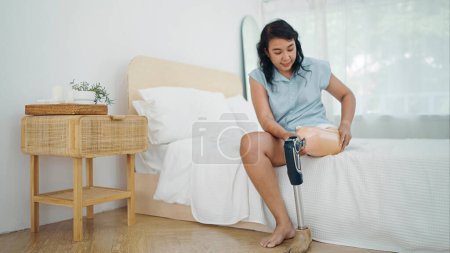 Téléchargez les photos : Adultes femmes asiatiques amputées enlever la jambe de la prothèse tout en étant assis sur le lit. Difficultés dans la vie des personnes handicapées, Handicap physique, Prothèse de jambe, Concept d'amputé - en image libre de droit