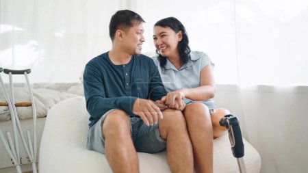 Photo for Happy asian woman in leg prosthesis and her husband sitting on comfortable sofa spending time together at home. Happy asian couple, Leg prosthetic equipment - Royalty Free Image