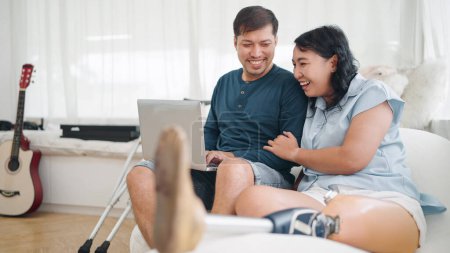 Téléchargez les photos : Heureuse femme asiatique dans la prothèse de jambe et son mari assis sur un canapé confortable tout en tapant sur ordinateur portable pour travailler en ligne ou bavarder sur les médias sociaux à la maison. Équipement de prothèse de jambe - en image libre de droit