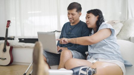 Photo for Happy asian woman in leg prosthesis and her husband sitting on comfortable sofa while typing on laptop for working online or chatting on social media at home. Leg prosthetic equipment - Royalty Free Image