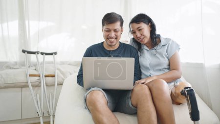 Photo for Happy asian woman in leg prosthesis and her husband sitting on comfortable sofa while typing on laptop for working online or chatting on social media at home. Leg prosthetic equipment - Royalty Free Image