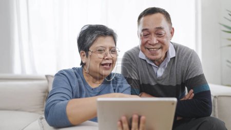 Téléchargez les photos : Heureux couple asiatique âgé parlant ensemble et en utilisant la tablette profiter de la navigation sur les médias sociaux tout en étant assis dans le salon à la maison. Couple de personnes âgées amour moment. Couple retraite style de vie - en image libre de droit
