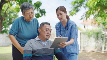 Foto de Asian woman nurse or caregiver showing treatment effect on tablet to elderly male patient sitting on wheelchair and his wife while relaxing at outdoors - Imagen libre de derechos