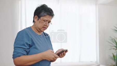 Téléchargez les photos : Asiatique femme d'âge moyen portant des lunettes tout en utilisant smartphone textos sur la navigation sur les médias sociaux ou des achats en ligne à la maison. Concept de retraite senior - en image libre de droit