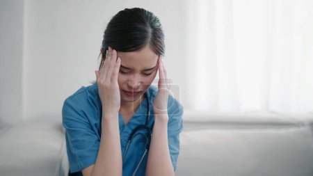 Foto de Cansada deprimida joven asiática enfermera en uniforme sufren de dolor de cabeza después de trabajar duro. Mujer triste agotada médico siente el estrés por agotamiento. Burnout médico - Imagen libre de derechos