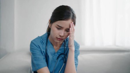 Téléchargez les photos : Fatigué déprimé jeune asiatique infirmière en uniforme souffrent de maux de tête après avoir travaillé dur. Épuisé femme triste médecin se sent le stress burnout. Épuisement professionnel du médecin - en image libre de droit