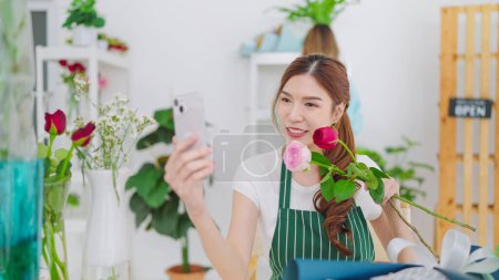 Téléchargez les photos : Jeunes femmes asiatiques propriétaire magasin floral tenant des fleurs roses sur les mains montrant au client en choisissant par appel vidéo sur smartphone pour prendre commande. Concept de petite entreprise - en image libre de droit