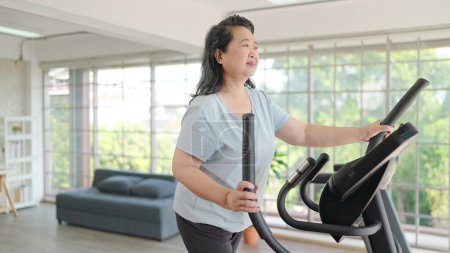 Téléchargez les photos : Femmes âgées asiatiques faisant de l'exercice sur la machine dans le salon à la maison. Femme mûre aiment faire de l'exercice sur tapis roulant pour le bien-être amincissant poids. Concept de mode de vie âgé - en image libre de droit