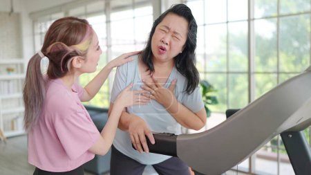 Téléchargez les photos : Les femmes âgées asiatiques ont des douleurs thoraciques pendant l'exercice sur la machine. Les jeunes femmes prennent soin de sa mère qui souffre de douleurs thoraciques en faisant de l'exercice. Concept de soins de santé - en image libre de droit