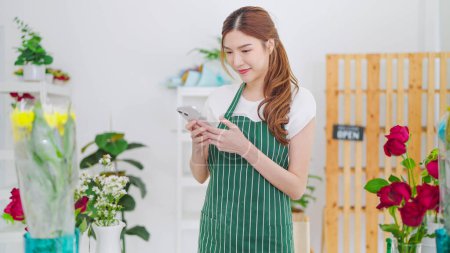 Photo for Beautiful young asian women owner floral store in apron typing on smartphone taking order with client or post on social media. Small business concept - Royalty Free Image