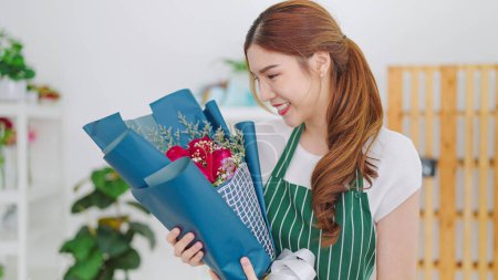 Téléchargez les photos : Belle jeune asiatique femme propriétaire magasin floral dans tablier tenant bouquet de fleurs de rose sur les mains passant bouquet au client à la boutique de fleurs. Concept de boutique de fleurs - en image libre de droit