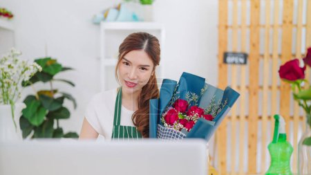 Téléchargez les photos : Jeunes femmes asiatiques propriétaire magasin floral tenant bouquet de fleurs roses sur les mains tandis que l'appel vidéo sur ordinateur portable communiquant avec le client. Vente bouquet en ligne en direct sur ordinateur portable - en image libre de droit