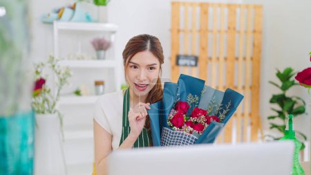 Téléchargez les photos : Jeunes femmes asiatiques propriétaire magasin floral tenant bouquet de fleurs roses sur les mains tandis que l'appel vidéo sur ordinateur portable communiquant avec le client. Vente bouquet en ligne en direct sur ordinateur portable - en image libre de droit