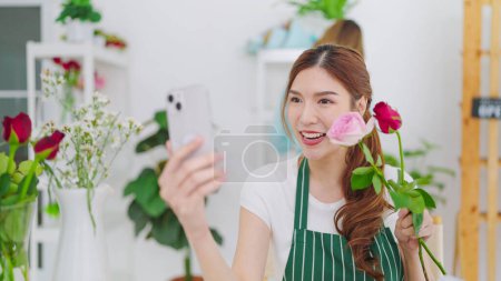 Photo for Young asian women owner floral store holding rose flowers on hands showing to client choosing through video call on smartphone for taking order. Small business concept - Royalty Free Image