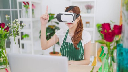 Foto de Joven asiática propietaria de una tienda floral con gafas VR moviendo las manos en el aire para trabajar y seleccionar flores a través de auriculares de realidad virtual. Concepto de negocio y tecnología, Metaverse virtual - Imagen libre de derechos