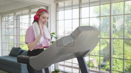 Photo for Happy young asian women in headphones waving hands, talking through video call on smartphone while exercising on the machine at home. Health care concept - Royalty Free Image