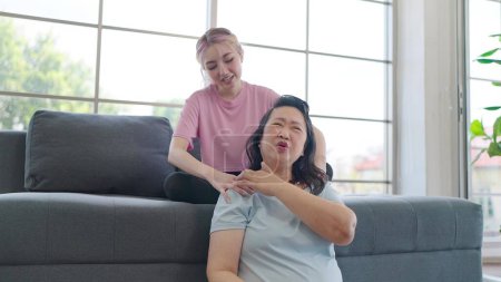 Téléchargez les photos : Fille asiatique donnant massage des épaules à la mère âgée tout en relexing dans le salon à la maison. Jeune fille donnant une mère âgée massage relaxant. Style de vie familial - en image libre de droit