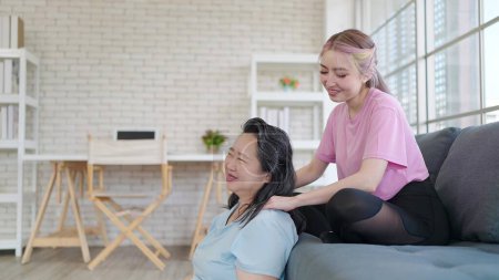 Téléchargez les photos : Fille asiatique donnant massage des épaules à la mère âgée tout en relexing dans le salon à la maison. Jeune fille donnant une mère âgée massage relaxant. Style de vie familial - en image libre de droit