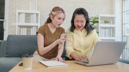 Photo for Young asian daughter teaching elderly mother using laptop sitting on couch in living room at home. Mother and daughter using laptop together. Learning how to use modern technologies - Royalty Free Image