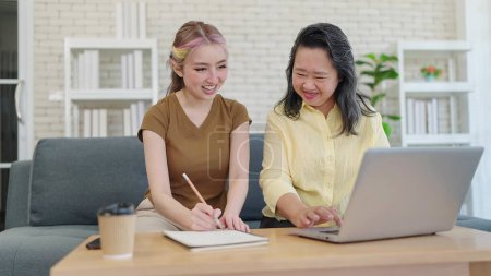 Photo for Elderly Asian mother and young daughter using laptop computer together in living room at home. Mother and daughter shopping through e-commerce website on laptop and taking note in notebook - Royalty Free Image